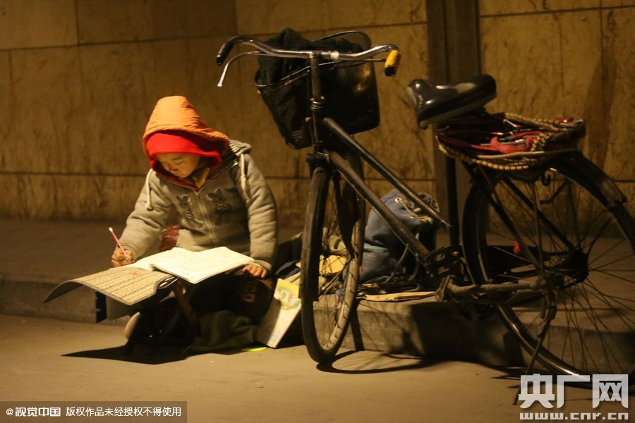 鄭州中原路京廣鐵路大橋下涵洞的馬路牙子邊，一位戴著紅色辣椒帽的小男孩趴在一個小馬扎上，上面鋪著一張車墊，在微弱的燈光下寫作業(yè)